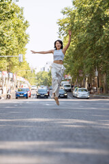 Obraz premium Young woman is happily jumping in the middle of a street with cars approaching in the background