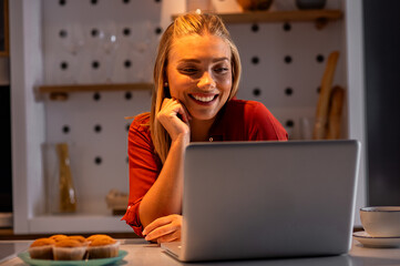 Smiling woman at home in kitchen using laptop while drinking tea at night.