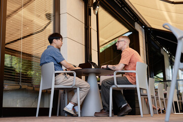 Fototapeta premium Side view of two adult male IT programmers using laptops to write code discussing work strategy while sitting at table in outdoor seating area of cafe, copy space