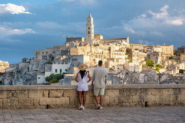 A couple stands on a stone balcony, gazing at the breathtaking skyline of Matera. The ancient...