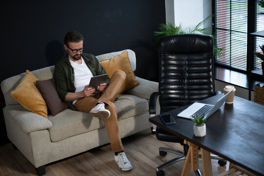 A man is sitting on a couch with a tablet in his lap. He is wearing glasses and a white shirt. The room has a black leather chair and a desk with a laptop and a cell phone