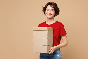 Young smiling happy satisfied cheerful woman she wear red t-shirt casual clothes hold stack cardboard blank boxes look camera isolated on plain pastel light beige background studio. Lifestyle concept.