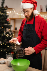 Young bearded man in apron and santa hat making dough for christmas gingerbread cookies in the kitchen, happy guy cooking pouring flour during winter holidays.