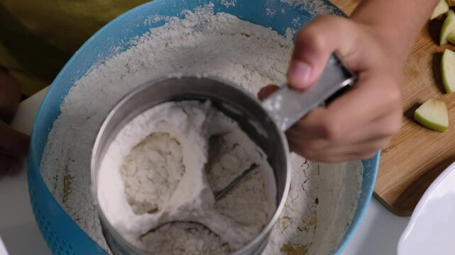 person sifts flour using a flour sifter into a bowl while preparing ingredients for baking. This image highlights a key step in baking, emphasizing the process of making homemade treats.