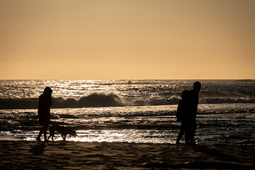 A faily walking a dog on the beach at sunset, in silhouette, warm orage summer sky with waves in the background, horizontal