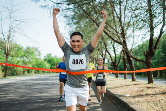 asian male runner crossing finish line celebrating victory with hands up gesture