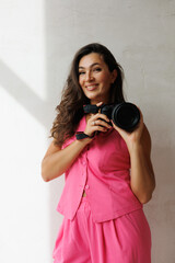 Obraz premium A beautiful tall brunette with spectacular styling stands at the window with a camera and smiles. A woman in a pink pantsuit in a vest and trousers holds a large camera and smiles. A photographer girl