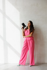 A beautiful tall brunette with spectacular styling stands at the window with a camera and smiles. A woman in a pink pantsuit in a vest and trousers holds a large camera and smiles. A photographer girl