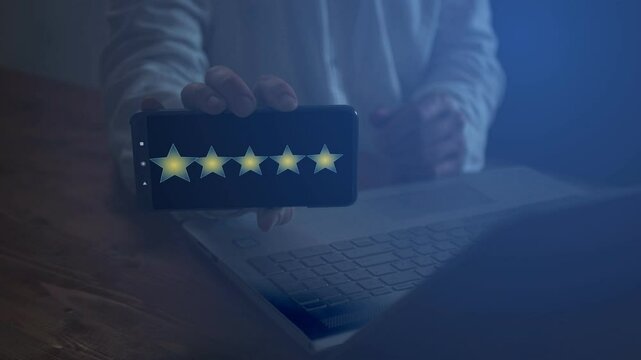 A man behind a laptop with a smartphone in his hand shows his screen in the dark on which there is footage with five stars that increase and decrease in turn. Victory and success concept. Cg