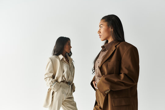 Two young and fashionable women pose confidently in stylish attire against a simple backdrop.