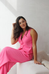 A beautiful tall brunette with spectacular styling stands at the window and smiles. A woman in a pink pantsuit in a vest and trousers on beige sofa. 