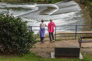 two women, one with a pink coat the other with a blue one, stand leaning on the rail looking over the weir that takes the River Aire past historic Salts Mill
