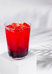 Red cocktail with ice and blackberries on white background with palm leaf shadow