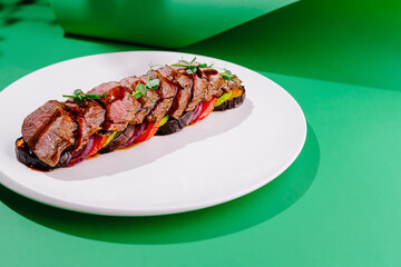 Sliced grilled beef with vegetables on white plate over green background