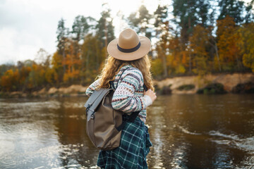 Female traveler with a backpack and a hat enjoying autumn hiking along the river. Active lifestyle....