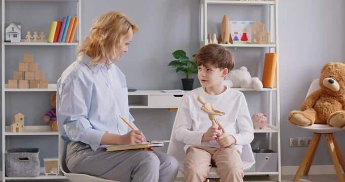 Young friendly professional female psychologist doctor consulting child boy patient during therapy session making notes in clipboard in office. Kids mental health concept. 4k video.