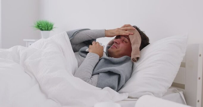 Sick young man lying in bed wrapped in blanket with wet towel on his head, suffering from fever, sneezing in tissue. Unhappy exhausted guy with cold, seasonal infection or flu virus feeling unhealthy.