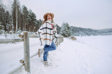 Fototapeta premium Portrait of a young smiling woman in hat, scarf, and sweater in winter time outdoors. Fashion. Enjoying nature. Boho style.
