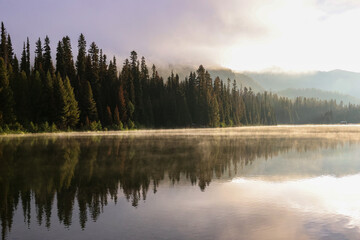 the steaming lightning lake in British Columbia in the early morning sunlight, while the forest is reflected in the calm water surface