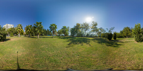 A large grassy park area under a clear blue sky with the sun shining brightly overhead. Palm trees and other vegetation create a peaceful, natural space in this 360-degree panorama.
