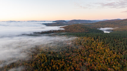Aerial view of a mountain range covered in vibrant fall foliage, with mist and fog drifting over the colorful forested hills, capturing the beauty of the autumn season.
