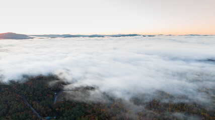 Aerial view of a sunrise over a forested mountain landscape, with dense mist and clouds creating a golden, ethereal atmosphere, showcasing the beauty of nature during early morning.