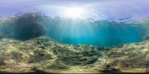 A sunlit underwater scene with large rock formations and a clear view of the seabed, captured in a 360-degree panorama. The bright sunlight creates a calm, immersive marine experience.
