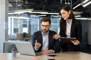 Colleagues in formal attire engaged in business meeting. Woman pointing at laptop screen, man...