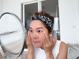 Asian Thai woman using moisturizer cream on face after shower in the morning, looking at mirror,...