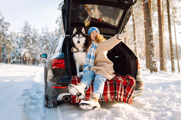 Young woman in winter clothes sitting in the trunk of a car with her husky and play. Winter holidays. Travel concept