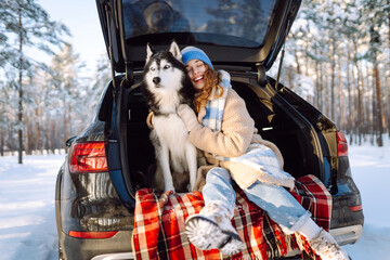 Young woman in winter clothes sitting in the trunk of a car with her husky and play. Winter holidays. Travel concept