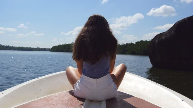 Unrecognizable girl with blowing long hair sits on bow of boat and admires a beautiful nature landscape. Young woman rests on deck of yacht and enjoys summer travel. Concept of vacation or holiday