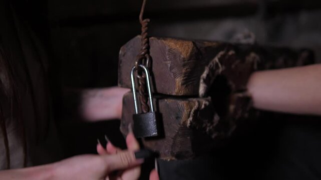 The girl closes the old rusty lock on the old wooden shackles. A scary quest room with creepy handcuffs. An old prison room for completing quests.