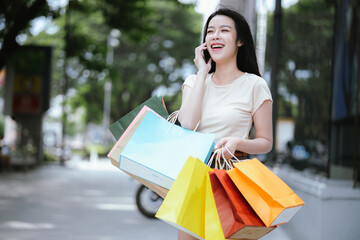 A modern young woman smiles as she walks in front of a shopping mall, each carrying shopping bags. One is checking her smartphone, enjoying the modern city life and shopping experience.