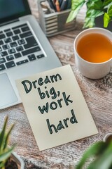 A handwritten note with the quote "Dream big, work hard" placed on a desk next to an open laptop and a cup of tea, in a modern workspace with a motivational vibe.