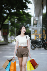 A cheerful young Asian woman, stylishly dressed, smiles excitedly while holding shopping bags....