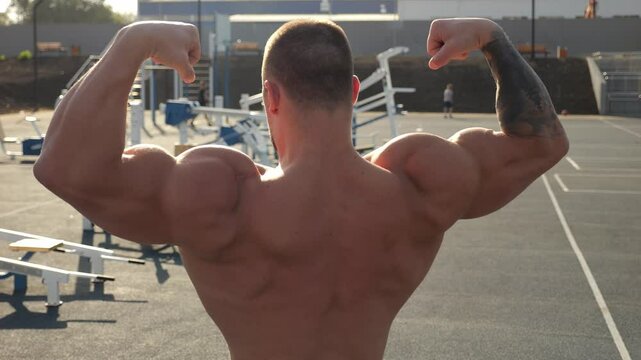 A handsome, muscular man shows off a toned back and arms. A bodybuilder shows off a toned body on an outdoor sports field at sunset. Outdoor exercise equipment. The image is stabilized.