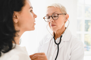 Serious caucasian female family doctor general practitioner listening in stethoscope female mature patient`s chest, heart beat at hospital appointment