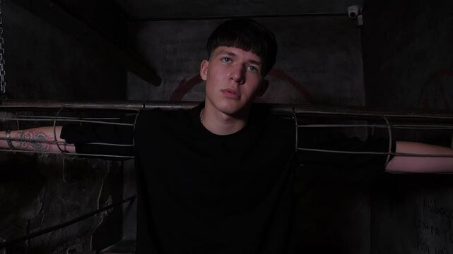 A young man is chained to horizontal handcuffs in a dark quest room. A dark prison room with old rusty walls and antique shackles. The room of horrors.