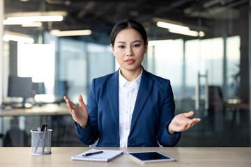 Asian business woman in office setting looking at camera, exhibiting serious demeanor while speaking, conveying professionalism and focus, accompanied by phone and notebook for notes.