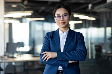 Asian business woman confidently standing in office, arms crossed, wearing glasses and suit....