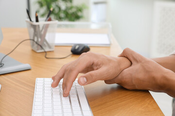 Man suffering from pain in wrist while working on computer at table indoors, closeup. Carpal tunnel...