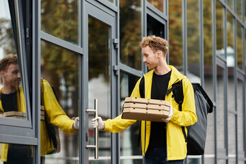 A delivery man carries multiple boxes as he enters an office building in daylight.