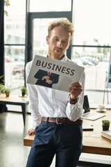 A redhead professional in a white shirt studies a business report while standing in a contemporary office.