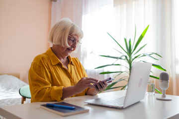 Focused old woman at home using laptop. Senior stylish entrepreneur with notebook and pen wearing...