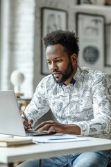 Focused man in a white shirt sitting at a desk writing notes while using a laptop in a modern office setting.