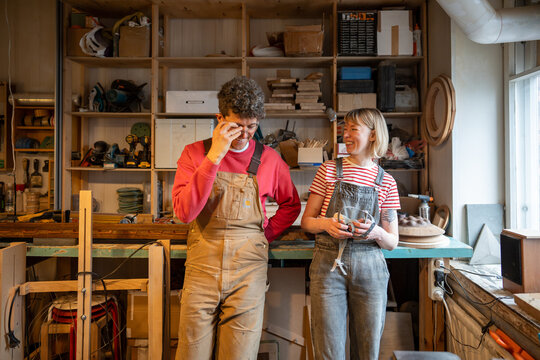 Cheerful carpenter couple laughs, goofs off during break in workshop. Family business, teamwork, shared passion, creative collaboration, small business, work life balance, joyful working partnership