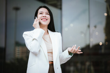 A successful Asian businesswoman stands smiling confidently in the city and plays with her mobile phone. She is professionally dressed and full of determination and ambition in the finance and investm