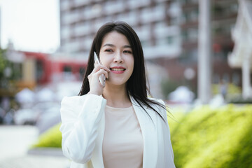 Fototapeta premium Active Asian woman in suit standing outdoors in city holding smartphone and laptop. She represents the future of finance and investment, keeping updated on news and opportunities.