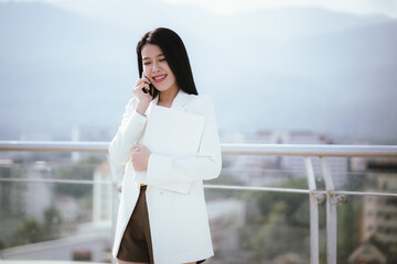 Fototapeta premium Confident Asian businesswoman walks through a bustling city smiling while using her mobile phone. She wears a suit and lives in the city, reflecting success and connections in the corporate world.